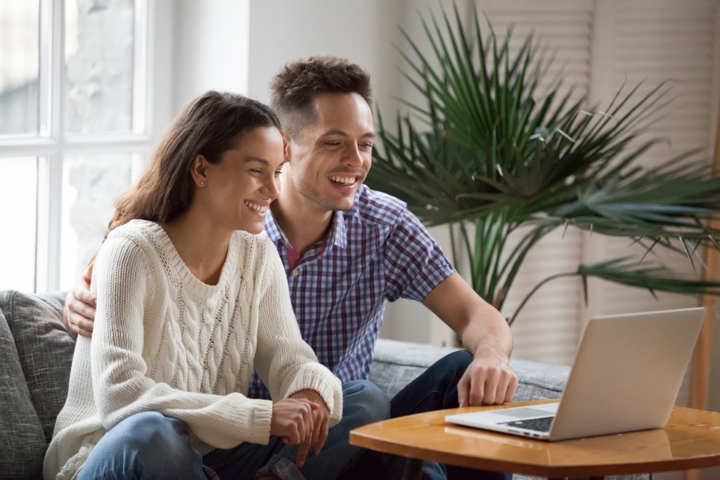 Happy young couple laughing watching funny video or comedy movie online, cheerful man and woman having fun enjoying videocall looking at laptop screen and smiling sitting on sofa at home together